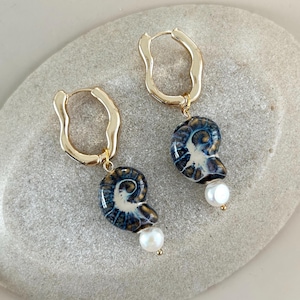 May include: A pair of gold-tone hoop earrings. Each earring features a blue and white ceramic pendant with a spiral design and a small white pearl. The earrings are displayed on a light gray stone surface.