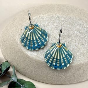 Op de afbeelding: Een paar schelpvormige oorbellen. De oorbellen zijn keramisch met een blauw en wit patroon en zilverkleurige metalen ringen. Ze worden gepresenteerd op een lichtgrijze stenen ondergrond. Groen gebladerte is zichtbaar.