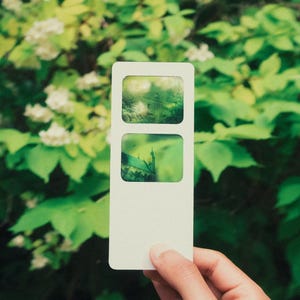 May include: A rectangular white card featuring two stacked rectangular cutouts. Each cutout displays a translucent, green-tinted photographic image of a building exterior, showing architectural details through the semi-transparent material.