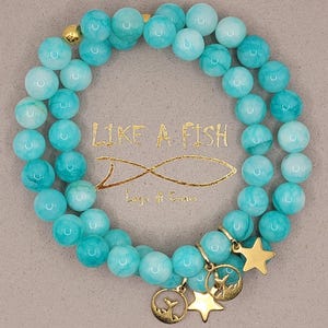 Può includere: Un braccialetto a doppia fila con perline rotonde turchesi. Ciondoli dorati tra cui una stella e due pendenti circolari con un design a coda di balena. Il testo "LIKE A FISH" e "Lago di Como" sono visibili.