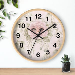 May include: A round wall clock with a light wood frame. The clock face features a pink floral design with a whimsical face in the center. Black hands indicate the time. The numbers are black and the clock has a light pink background.
