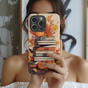 May include: A phone case with a fall-themed design featuring a stack of books, autumn leaves, and pumpkins. The case is cream-colored with a brown border.