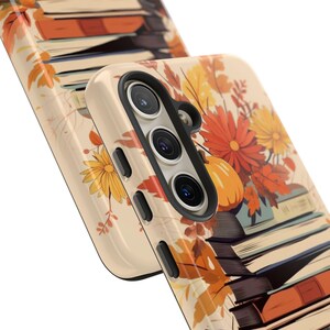 May include: A phone case with a fall-themed design featuring a stack of books, autumn leaves, and a pumpkin. The case is beige with a black camera cutout.