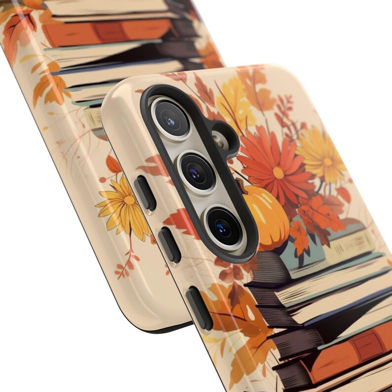 May include: A phone case with a fall-themed design featuring a stack of books, autumn leaves, and a pumpkin. The case is beige with a black camera cutout.