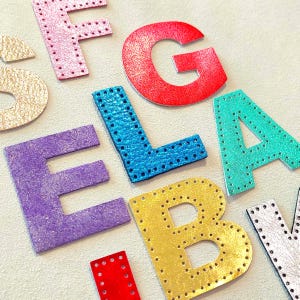 May include: Assorted colorful leather letters, including red, blue, purple, and gold, are arranged on a light background. Each letter is stitched around the edges. The letters spell out the name "ELA".