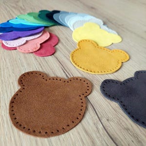 Puede incluir: Un conjunto de formas de oso de fieltro en varios colores, incluyendo marrón, amarillo y negro. Las formas están cosidas con una línea punteada decorativa alrededor de los bordes.