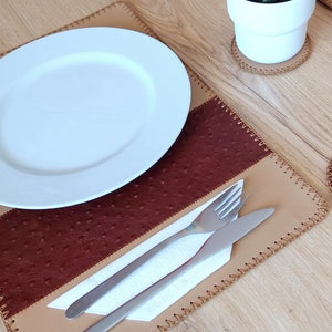 Könnte beinhalten: Ein braunes und beiges Leder-Platzset mit einem genähten Rand. Ein weißer Teller, eine Gabel, ein Messer und eine weiße Serviette liegen auf dem Platzset.