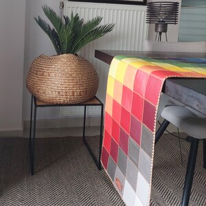 May include: A colorful patchwork table runner with a geometric design in shades of red, yellow, orange, and gray. The runner is made of leather and has a fringed edge. It is displayed on a wooden table with a woven rug underneath.