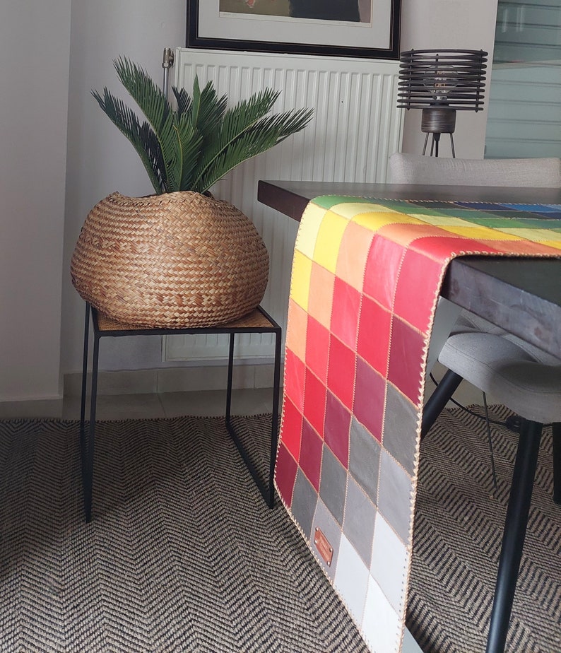May include: A colorful patchwork table runner with a geometric design in shades of red, yellow, orange, and gray. The runner is made of leather and has a fringed edge. It is displayed on a wooden table with a woven rug underneath.