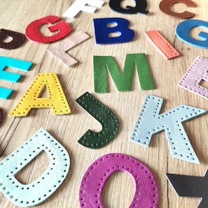 Op de afbeelding: Diverse kleurrijke leren letters, waaronder een rode 'G', een blauwe 'B' en een groene 'M', liggen op een houten oppervlak. Elke letter is met contrasterend garen met de hand gestikt. De letters variëren in grootte en lettertype, wat een decoratief arrangement creëert.