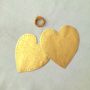 Può includere: Due forme di cuore in pelle dorata con bordi cuciti, pronte per l'artigianato. Una piccola bobina di filo dorato è sopra, suggerendo un progetto fai-da-te. I cuori sono su una superficie grigio chiaro.