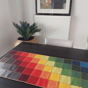 May include: A colorful table runner made of leather squares in shades of red, orange, yellow, green, blue, and gray. The runner is stitched together with a gold thread and has a fringe border.