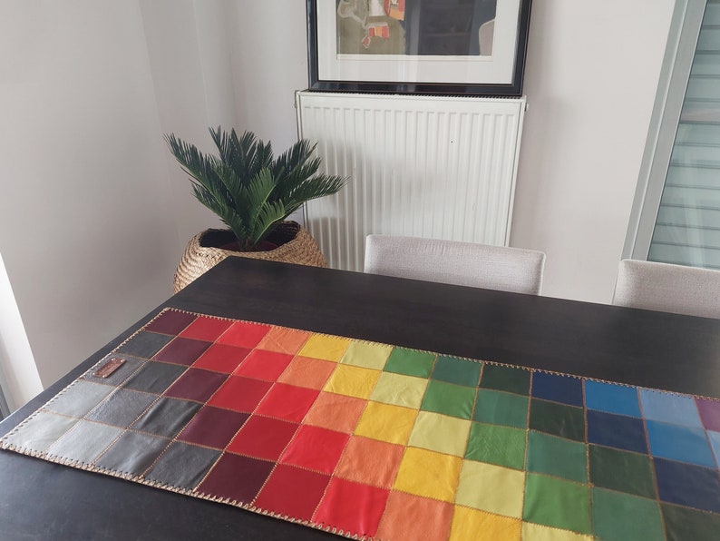 May include: A colorful table runner made of leather squares in shades of red, orange, yellow, green, blue, and gray. The runner is stitched together with a gold thread and has a fringe border.
