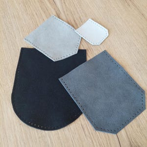 Puede incluir: Un conjunto de piezas de cuero en varias formas y colores, incluyendo negro, gris y gris claro. Cada pieza tiene un borde cosido con agujeros espaciados, lo que sugiere que son componentes para un proyecto de artesanía o marroquinería.