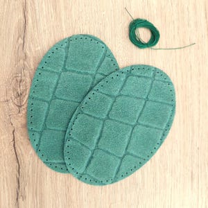Peut inclure: Deux pièces ovales en daim vert avec un motif quadrillé matelassé et des trous pré-perforés sur les bords pour la couture, accompagnées d'une petite bobine de fil vert assorti pour la fixation sur des vêtements ou autres textiles.