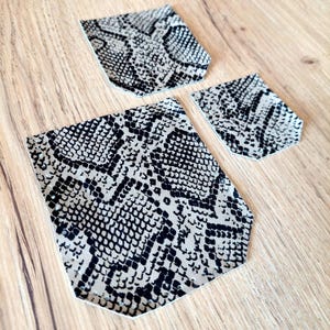 Op de afbeelding: Drie stukken imitatieleer met een slangenprint in zwart en beige. De stukken zijn in zakvormen gesneden, met één grote en twee kleinere maten. Het materiaal heeft een textuur.