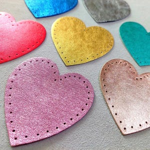 Peut inclure: Plusieurs patchs en forme de cœur en cuir, de couleurs variées : rouge, or, rose, bleu et turquoise. Chaque cœur présente une surface texturée et est bordé de petits trous espacés, probablement pour la couture. Les patchs sont disposés sur une surface gris clair.