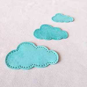 以下が含まれることがあります： ターコイズ色の雲の形をした革の切り抜きが3つ。縁はスカラップで、ステッチのディテールが施されています。雲のサイズは異なり、一番大きいものが一番下に、一番小さいものが一番上にあります。背景は明るいニュートラルカラーです。