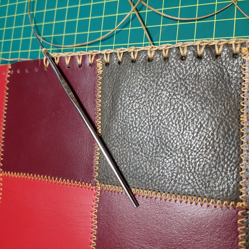 May include: A close-up of a leather patchwork project with a crochet hook. The leather is stitched together with a zig-zag stitch and the edges are finished with a brown crochet border.