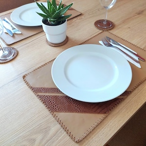 Könnte beinhalten: Ein braunes Leder-Platzset mit einem genähten Rand und einem dekorativen Muster. Ein weißer Teller, Besteck und ein Glas befinden sich auf dem Platzset.