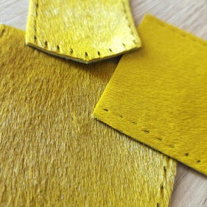 May include: Close-up of three pieces of yellow cowhide leather. The leather has a soft, furry texture and is a vibrant shade of yellow. Two pieces have stitched edges, suggesting they are cut for a specific purpose.