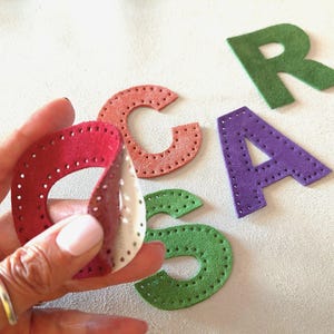 Może przedstawiać: Ręka trzymająca czerwoną, białą i bordową skórzaną literę. Litery G, A i R są rozrzucone w pobliżu. Litery są w odcieniach zieleni, fioletu i koralu. Każda litera ma szytą obwódkę i szereg małych otworów.