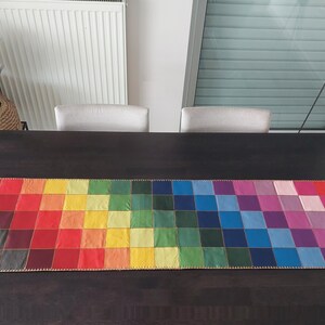 May include: A colorful table runner with a rainbow pattern. The runner is made of squares of different colors, including red, orange, yellow, green, blue, indigo, and violet. The runner is on a dark brown wooden table.