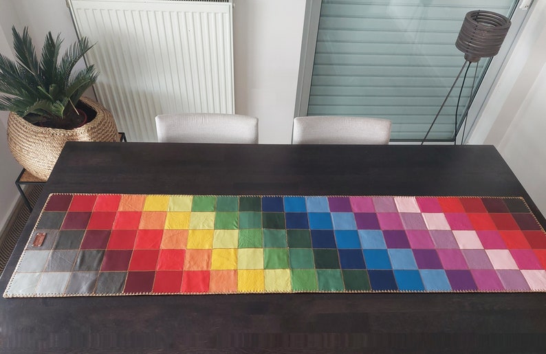 May include: A colorful table runner with a rainbow pattern. The runner is made of squares of different colors, including red, orange, yellow, green, blue, indigo, and violet. The runner is on a dark brown wooden table.