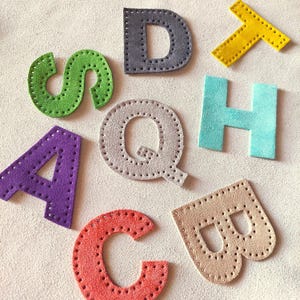 May include: A selection of colourful, stitched felt letters in various shades, including green, purple, grey, red, blue, yellow, and tan. The letters are arranged on a light beige surface, showcasing their individual shapes and textures. The letters are from the alphabet.