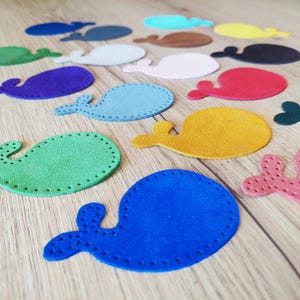 May include: A collection of felt whale shapes in various colours, including blue, green, red, yellow, and purple. The whales are all stitched with a decorative pattern around the edges.
