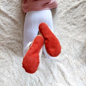 May include: A pair of soft slippers featuring bright orange suede soles with visible white whipstitching around the edges. The slippers have a plush, cream-colored interior lining and are worn over white leggings and a pink top.