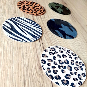 May include: Five round coasters featuring various animal print designs. The coasters are crafted from a brown material and have a white background. The designs include zebra stripes, leopard spots, and camouflage patterns.
