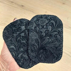 May include: Two black suede patches with a floral design. The patches have a decorative stitched edge.