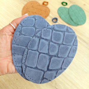 May include: Two oval-shaped, blue suede insoles with a textured, crocodile-like pattern. Other suede shapes are in the background, including a heart and oval shapes in brown and green. The insoles are held in a hand.