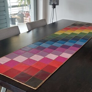 May include: A colorful leather table runner with a checkered pattern in shades of pink, red, purple, blue, and green. The runner is made of leather squares sewn together with a gold thread.
