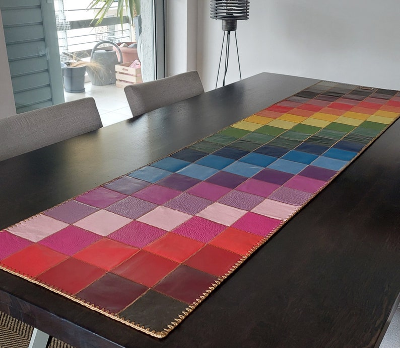 May include: A colorful leather table runner with a checkered pattern in shades of pink, red, purple, blue, and green. The runner is made of leather squares sewn together with a gold thread.