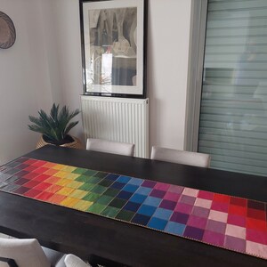 May include: A rainbow checkered table runner with a gold trim. The runner is on a dark wood table with two chairs.