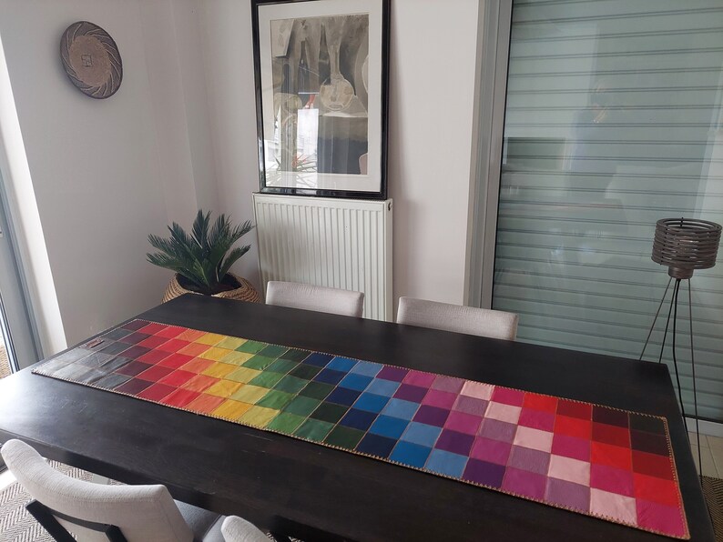 May include: A rainbow checkered table runner with a gold trim. The runner is on a dark wood table with two chairs.
