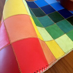 May include: A colorful patchwork leather placemat with a rainbow pattern of yellow, orange, red, green, blue, and light blue squares. The placemat is folded in half, showing the stitching along the edges.
