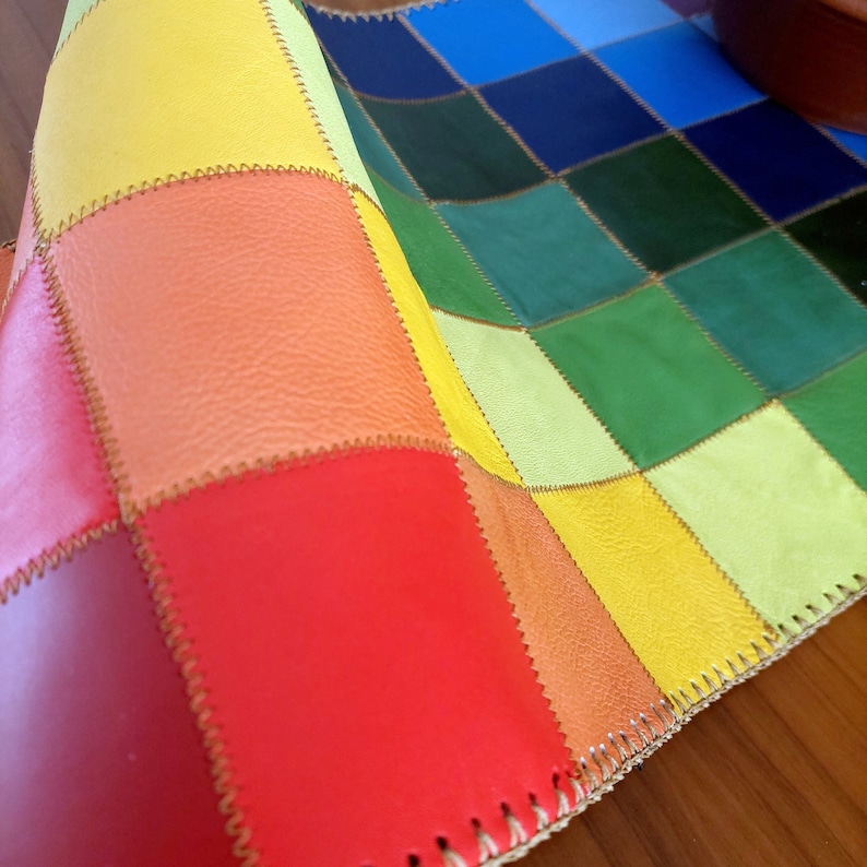 May include: A colorful patchwork leather placemat with a rainbow pattern of yellow, orange, red, green, blue, and light blue squares. The placemat is folded in half, showing the stitching along the edges.