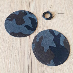 Puede incluir: Dos parches redondos de tela de camuflaje gris y negro con costuras negras alrededor de los bordes. También se ve una bobina de hilo negro.