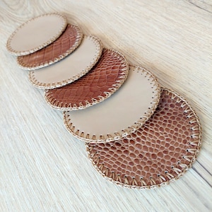 May include: Set of six brown and beige leather coasters with a stitched edge. The coasters have a textured, reptile-like pattern.