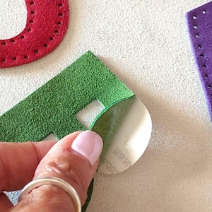 Könnte beinhalten: Nahaufnahme einer Hand, die eine weiße Kleberückseite von einem grünen Wildlederbuchstaben abzieht. Weitere Wildlederbuchenstaben in Rot und Lila sind sichtbar. Die Buchstaben haben genähte Kanten, wahrscheinlich für Bastel- oder DIY-Projekte.