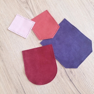 Puede incluir: Un surtido de piezas de cuero de gamuza de colores en varias formas. Las piezas son en tonos de rosa, rojo y morado, con bordes cosidos. Los artículos están dispuestos sobre una superficie de madera clara.