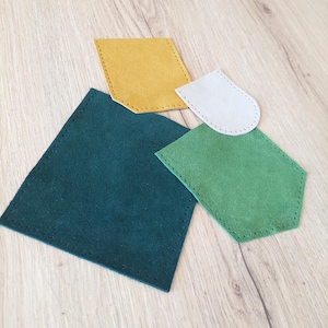 Op de afbeelding: Vier stukken suède leer in verschillende vormen en kleuren: teal, geel, groen en grijs. Elk stuk heeft een gestikte rand, mogelijk voor een knutselproject. Geplaatst op een houten oppervlak.