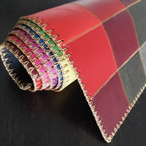 May include: A multi-colored leather placemat with a stitched edge. The placemat is rolled up and the different colors of leather are visible. The placemat is made of different shades of red, purple, gray, and black leather.