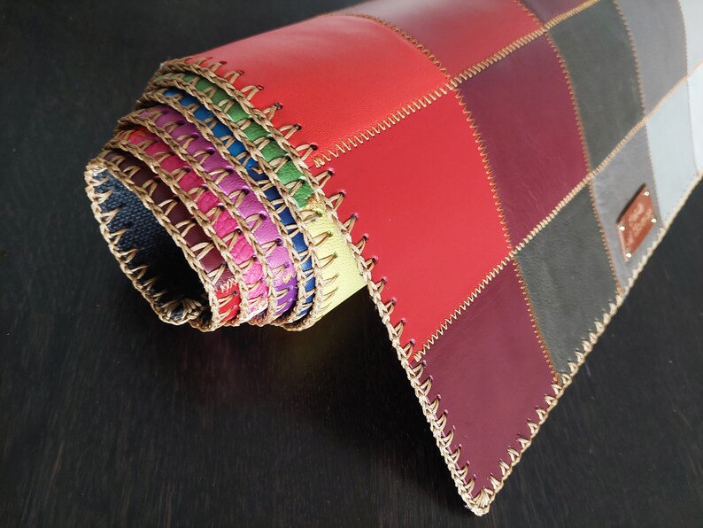 May include: A multi-colored leather placemat with a stitched edge. The placemat is rolled up and the different colors of leather are visible. The placemat is made of different shades of red, purple, gray, and black leather.