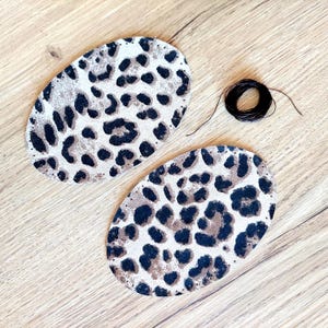 Puede incluir: Dos parches de cuero ovalados con un diseño de estampado de leopardo. Los parches tienen agujeros alrededor de los bordes y están listos para ser cosidos juntos. Se incluye un carrete de hilo negro.