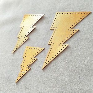 May include: Four gold-colored leather lightning bolt shapes with small, evenly spaced holes along the edges. The bolts vary in size and are arranged on a light gray surface. The leather has a slight sheen.