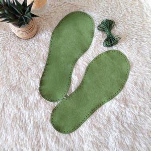Op de afbeelding: Een paar olijfgroene suède schoenzolen met voorgeponste gaten langs de randen om te naaien, vergezeld van een klein bundeltje bijpassend groen gewaxt garen voor montage, gepresenteerd als een knutselset voor het maken van schoenen.
